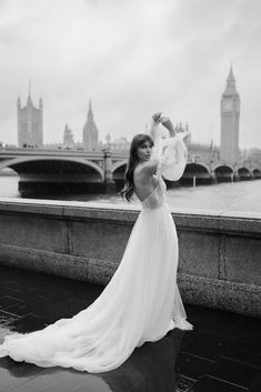 a woman in a long white dress standing on the side of a river with big ben in the background