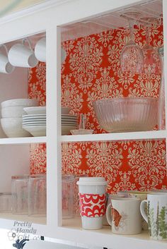 the shelves are filled with dishes, cups and other kitchen items in front of a red wallpapered background