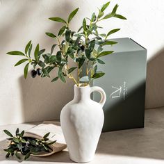 a white vase sitting on top of a table next to a plate with an olive branch in it