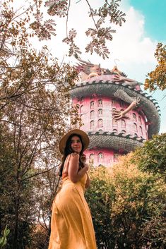 a woman in a yellow dress and hat posing for the camera with a pink building behind her