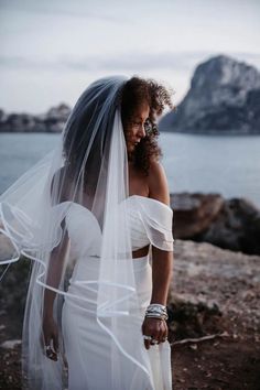 a woman in a wedding dress is standing by the water with her veil over her head