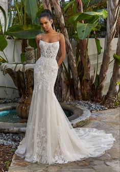 a woman in a wedding dress is standing near a fountain and palm trees with her hand on her hip