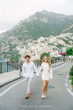 a man and woman holding hands walking down the road in front of a mountain town