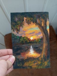 a person holding up a painting of a sunset over a lake with trees in the foreground