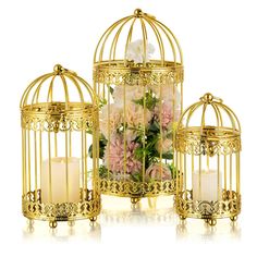 three gold birdcages with flowers and candles in them on a white table top