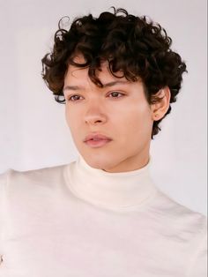 a young man with curly hair wearing a white turtle neck shirt and looking at the camera