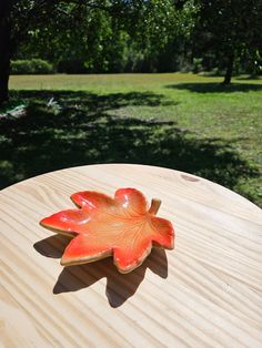 an orange leaf shaped dish sitting on top of a wooden table next to trees and grass