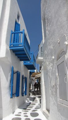 two people walking down an alley way with blue balconies and white buildings on either side