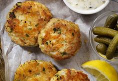 three crab cakes with pickles and lemon wedges next to them on a piece of wax paper