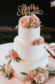 a three tiered wedding cake with flowers on the side and an acrylic name sign