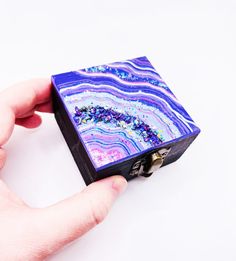 a hand holding a small purple and blue marbled box with metal latch on it