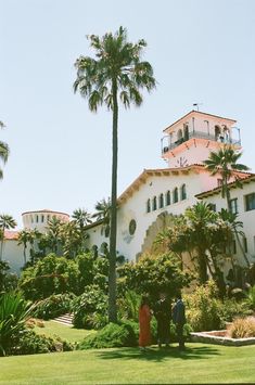 two people standing in front of a large white building with palm trees on the lawn