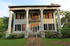 an old white house with columns and shutters on the front porch is surrounded by greenery