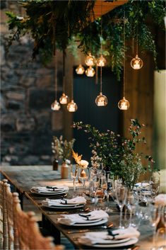 a dining table set with place settings and hanging lights over the dinner table is surrounded by greenery