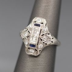 Elegant and classically geometrical, this Art Deco engagement ring features gorgeous old cut diamonds and sapphires. The double center stone design is unique with two emerald cut diamonds, bezel set and stacked on each other. The stones measure 3.5mm x 3mm, calculating to .20ct each. The diamonds are a VS clarity and H/I color. The stack is capped on top and bottom with French cut sapphires. The ring also features additional Old European cut diamonds on the top, bottom and sides. There are eight Diamond And Sapphire Ring, Art Deco Emerald, French Cut, Art Deco Engagement, Deco Engagement Ring, Art Deco Engagement Ring, European Cut Diamonds, Stone Design, Gorgeous Art