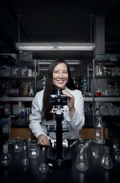 a woman standing in front of a microscope with lots of glass beakers around her