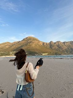 a woman is standing on the beach with her cell phone in her hand and mountains in the background