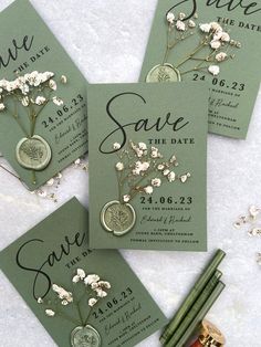 save the date cards with white flowers and coins on them, sitting next to some pencils