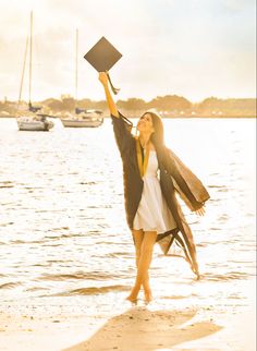 a woman holding up a graduation cap and gown on the beach with boats in the background