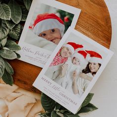 two christmas cards sitting on top of a wooden table next to green leaves and greenery