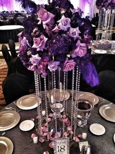 the table is set with silver plates and purple flowers in tall vases on each side