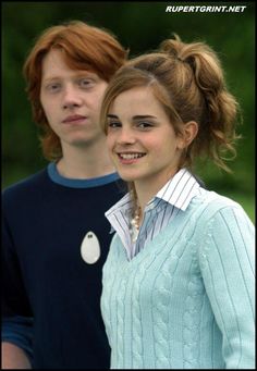 two young people standing next to each other in front of green trees and bushes, one wearing a blue sweater