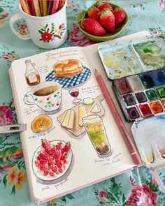 an open notebook with watercolors and food on it next to a bowl of strawberries