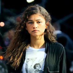 a woman with long hair is walking down the street wearing a t - shirt and jacket