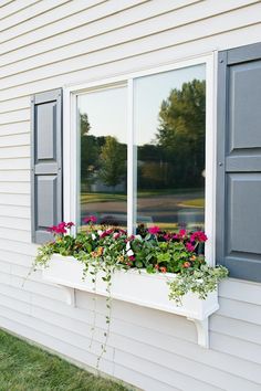 two windows with flowers in them on the side of a white house near grass and trees