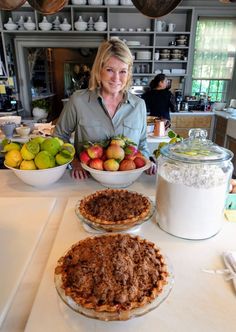 a woman standing behind two pies on top of a counter next to bowls of fruit