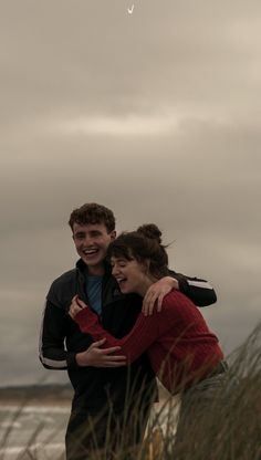 two people hugging each other in front of an overcast sky and the ocean behind them