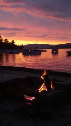 a campfire is lit up on the beach at sunset with boats in the water