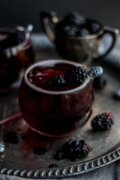 a glass filled with liquid and blackberries on top of a metal tray next to silver spoons