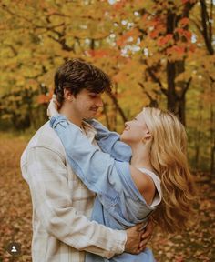 a man and woman standing in front of trees with leaves on the ground, looking into each other's eyes