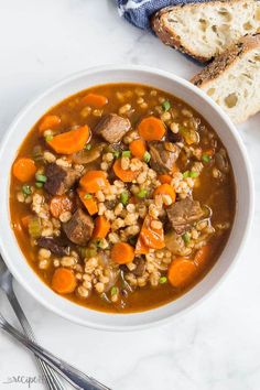 a bowl of beef barley soup with carrots, peas and bread on the side