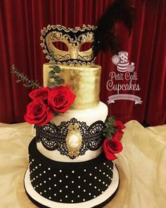 a three tiered cake with red roses and a mask on top, sitting on a table