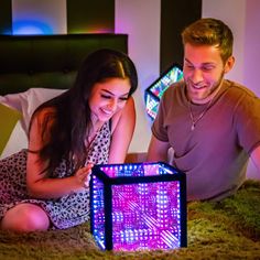 a man and woman sitting on the floor looking at a colorful light box in front of them