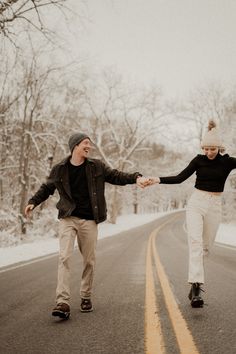 a man and woman holding hands while walking down the road in the snow with trees behind them