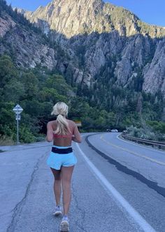 a woman running down the road in front of some mountains and trees with her back to the camera