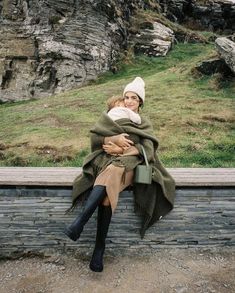 a woman holding a baby wrapped in a blanket sitting on top of a stone wall