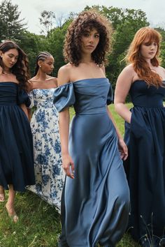 three women in dresses standing on grass with trees in the background and one woman wearing a blue dress