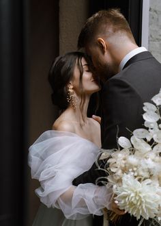 a bride and groom kissing in front of a window with white flowers on the windowsill