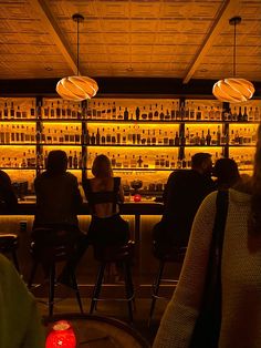 people sitting at tables in front of a bar with bottles on the shelves and lights hanging from the ceiling