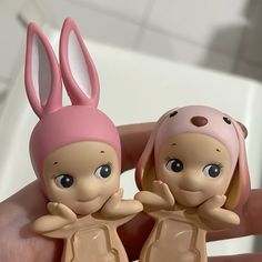 two little dolls are sitting in the palm of someone's hand, one is wearing a pink bunny hat