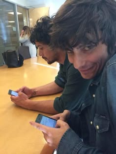 two young men sitting at a table looking at their cell phones while one looks at the camera