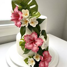 a three tiered cake with pink and white flowers on the top, next to a window