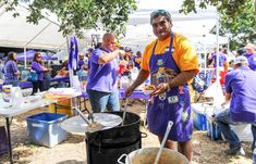 two men in purple and yellow aprons are cooking food at an outdoor event with other people