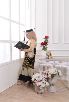 a woman in a costume reading a book next to flowers and a table with vases on it