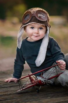 a small child wearing a hat and goggles sitting on the ground with a toy airplane