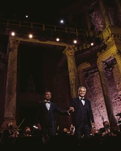 two men in tuxedos standing on stage with conductor and orchestra members behind them